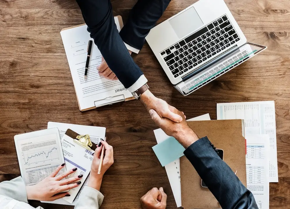 Two people shake hands over a wooden table laden with documents, a laptop, and charts, evoking the image of an accountants meeting. The papers show graphs and contractual details, with one person holding a pen. This scene captures the essence of business collaborations in Bromborough, Wirral.