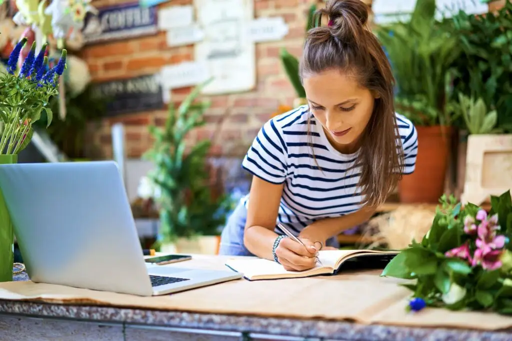 A woman in a striped shirt is writing in a notebook at a flower shop in Bromborough. She stands behind a counter with a laptop, surrounded by various plants and flowers. The background shows a brick wall with decorative signs, lending the Wirral shop an inviting charm.