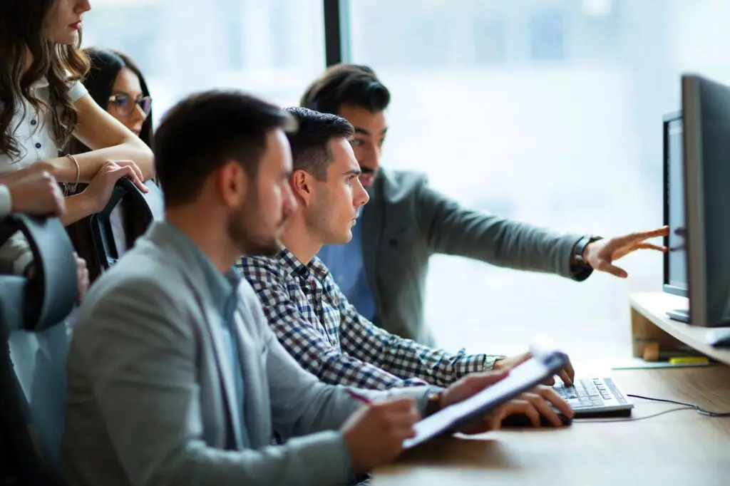 In an office near Birkenhead, a group of accountants gather around desks, focusing intently on computer screens. One person points at the monitor while others watch attentively, dressed in business attire—indicative of a focused work meeting or collaboration.