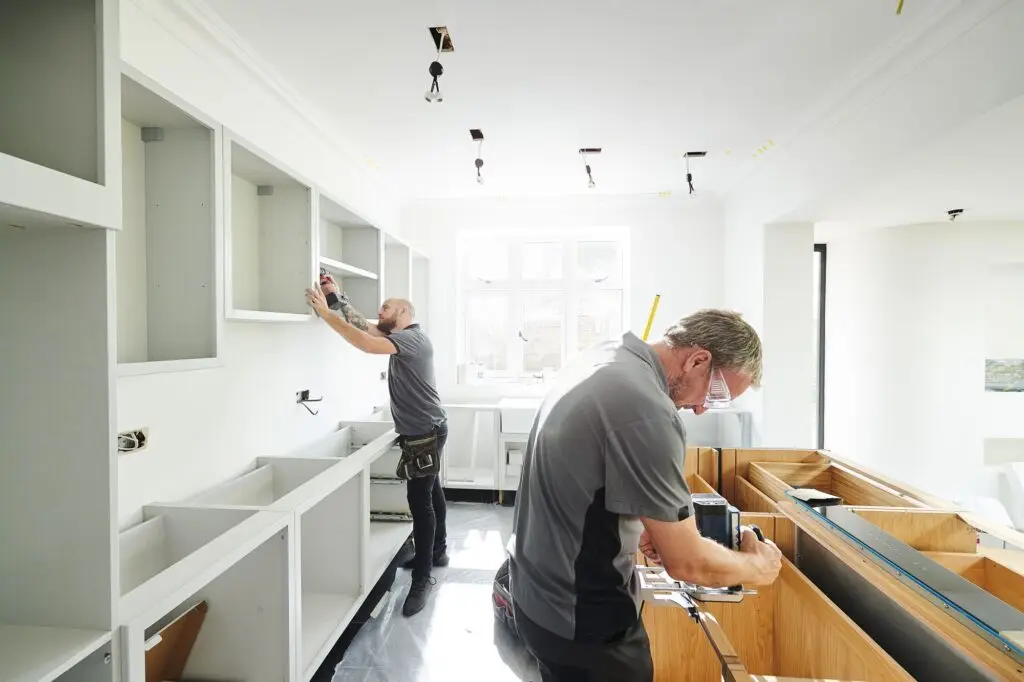 Two men are working in a bright kitchen under renovation in Birkenhead. One is installing an upper cabinet, while the other is measuring a countertop. The room is lit by a large window, and various tools are scattered around the space.