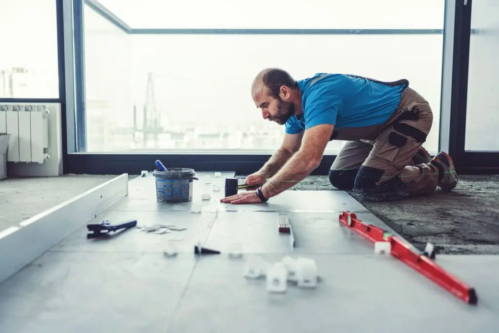 A man kneels on the floor, meticulously aligning tiles in a room with large windows. Dressed in a blue shirt and beige work pants, he works with precision. Nearby are tools, a bucket, and a level. Through the windows, views as orderly as an accountants ledger unfold across the industrial landscape.
