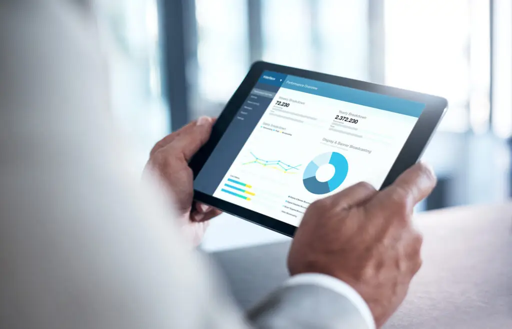 A person is holding a tablet showcasing analytics data, including a pie chart, line graph, and numerical figures against a blurred Birkenhead office background.