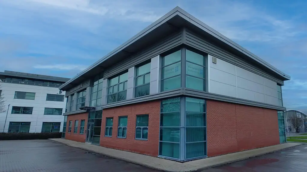 A modern, two-story office building with large windows and a flat roof, ideal for accountants. The bottom half is red brick, while the top gleams in metallic silver under a cloudy sky with patches of blue. A tidy path runs alongside the building, reflecting its professional appeal.