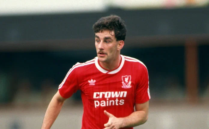 Soccer player in a red uniform with Crown Paints logo, running on a field during a match. The player has short dark hair and is focused on the game. Spectators are visible in the blurred background.